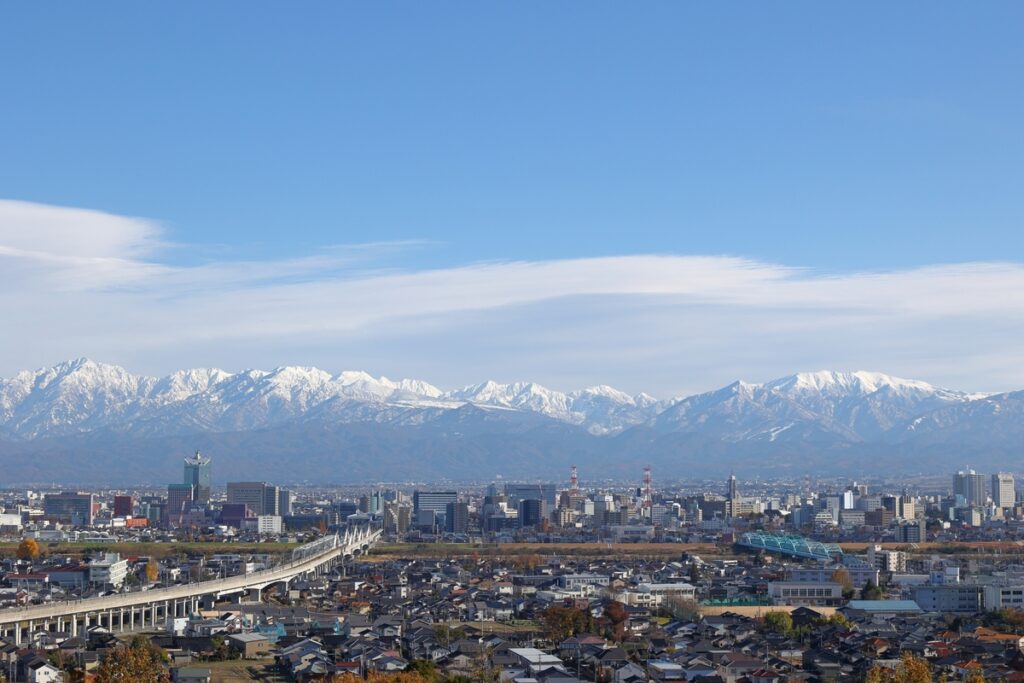 青空の下に広がる、雪を頂いた壮大な立山連峰を背景にした富山市街地の街並み