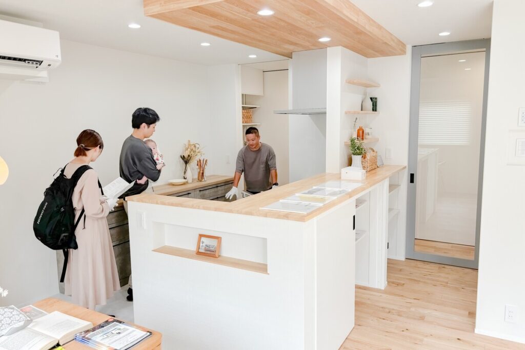 完成した新築住宅のキッチンで、引き渡し前の説明を受けるご家族。白を基調とした清潔感のあるキッチンカウンターや下がり天井の木目など、細部までこだわりが詰まった住まいの仕上がりを確認しているシーンです。