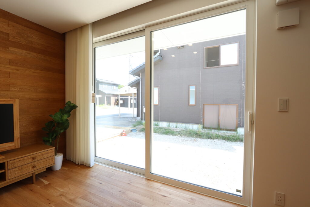 無垢フローリングと木目調のアクセントウォールを組み合わせたリビング。大きな引き違いの掃き出し窓からは外光が豊かに降り注ぎ、テレビボードなどの家具と調和した温かみのある内装です。