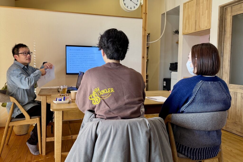 スクリーンに映し出された「家づくりの疑問点」を見ながら、工務店の担当者から住宅ローンや土地選びについての専門的なアドバイスを熱心に聞く夫婦の背面ショット。
