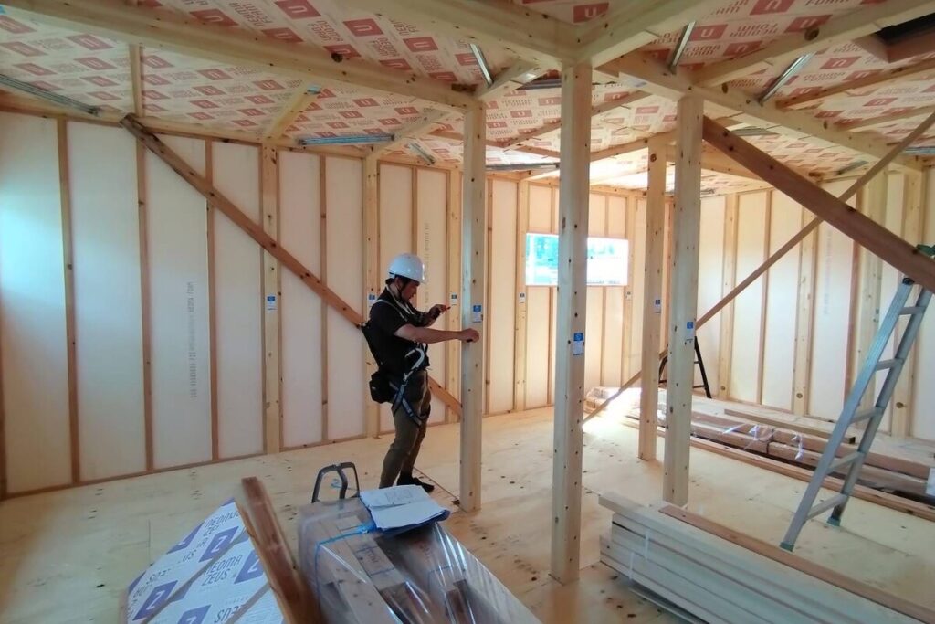 ヘルメットを着用した施工管理者が、建設途中の住宅内部で柱の垂直や精度を確認している現場風景。天井や壁には高性能断熱材が施工されており、安全と品質を徹底する工務店の家づくりの姿勢を捉えています。