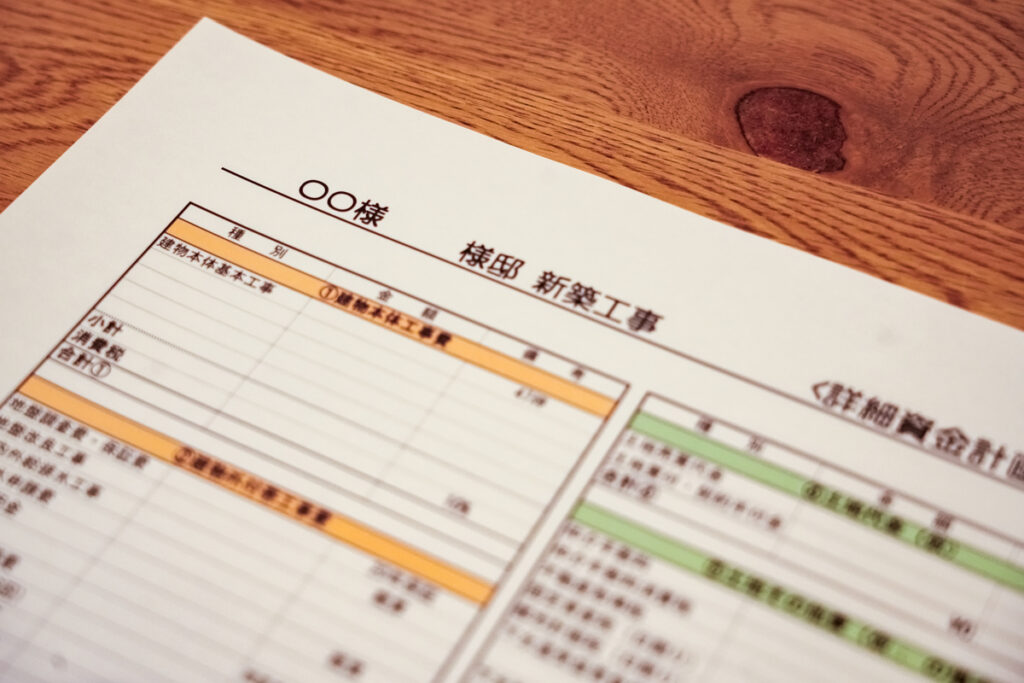 木目調のテーブルの上に置かれた「新築工事 詳細資金計画書」の書類。建物本体工事費や付帯工事費などの項目が並び、家づくりにおける予算計画や資金シミュレーションを行っている様子を表しています。