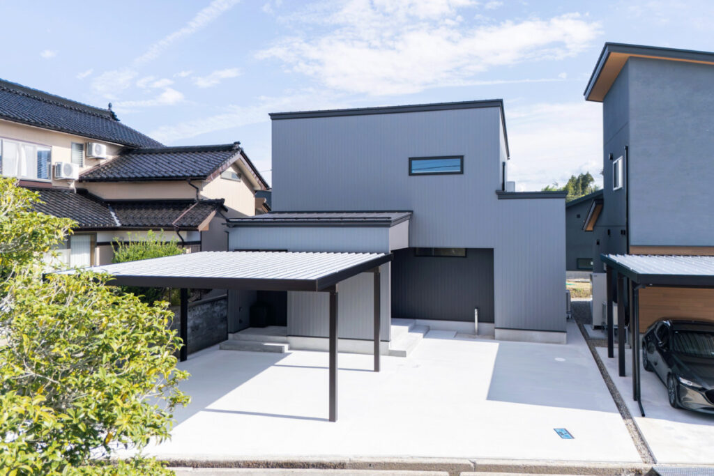 グレーのガルバリウム外壁とスクエアなフォルムが特徴的な、カーポートを備えたモダンなデザインの住宅。