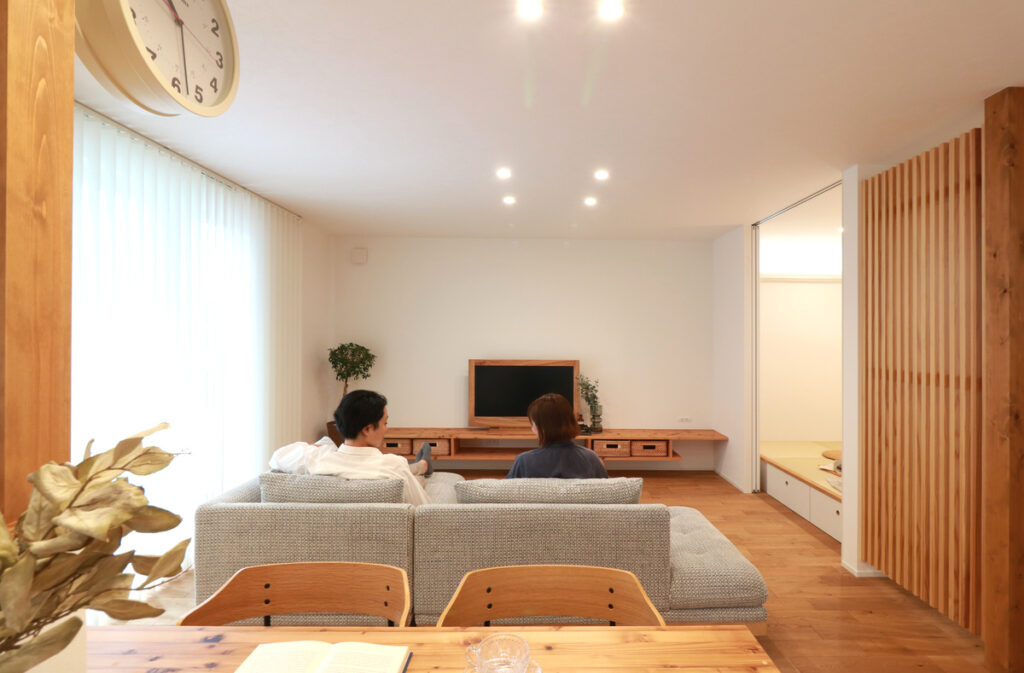 木の温もりを感じる造作テレビボードや格子パーテーションに囲まれた、開放感のあるナチュラルモダンなリビングでくつろぐ夫婦。