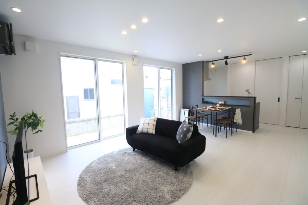 白を基調とした内装に黒のソファが映える、シンプルでスタイリッシュなモノトーンインテリアのリビング。奥にはキッチンとダイニングが広がり、掃き出し窓から明るい光が差し込んでいます。