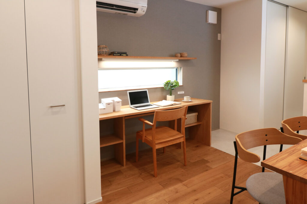 グレーのアクセントクロスを背景に、窓からの採光を活かした造作デスクと、北欧風の木製チェアを組み合わせたワークスペース。