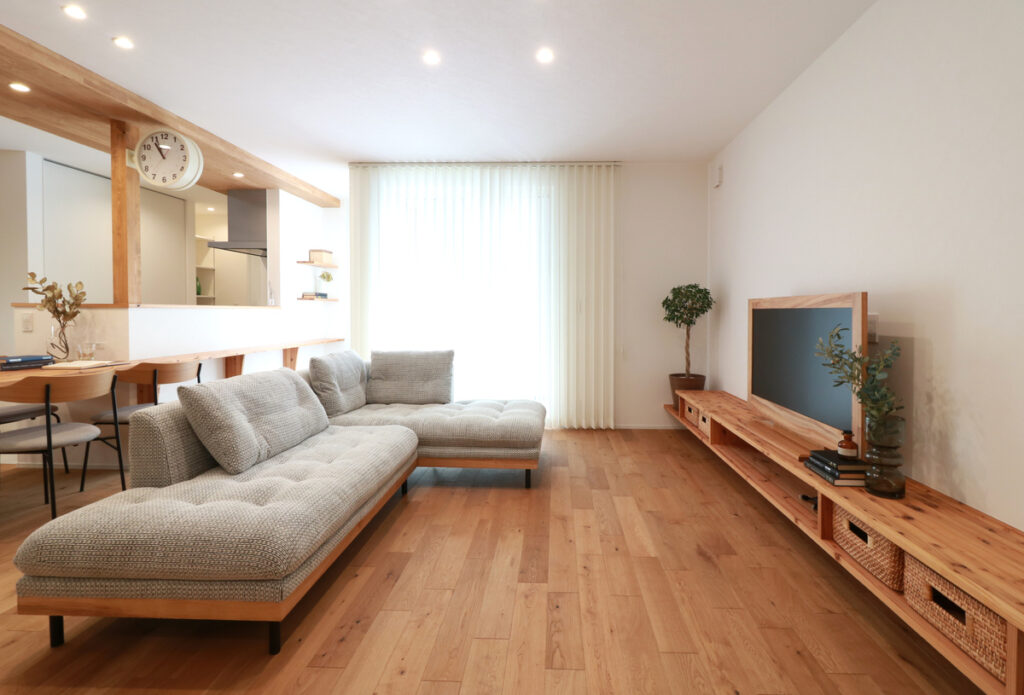 無垢材の床と白い壁に囲まれた、開放感あふれるナチュラルモダンなリビング。木の質感を生かした造作のロングテレビボードや対面キッチンが、明るい光が差し込む空間に心地よく調和しています。