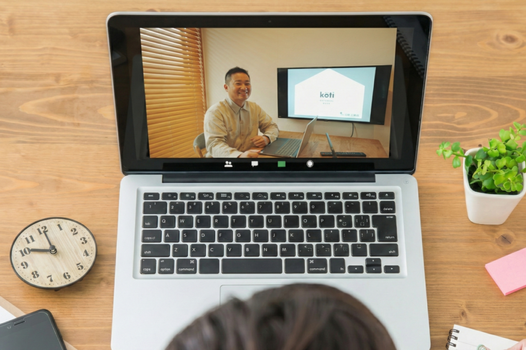 オンライン相談の画面に映る、木目調の壁とモニターを背景にした工務店スタッフの男性。