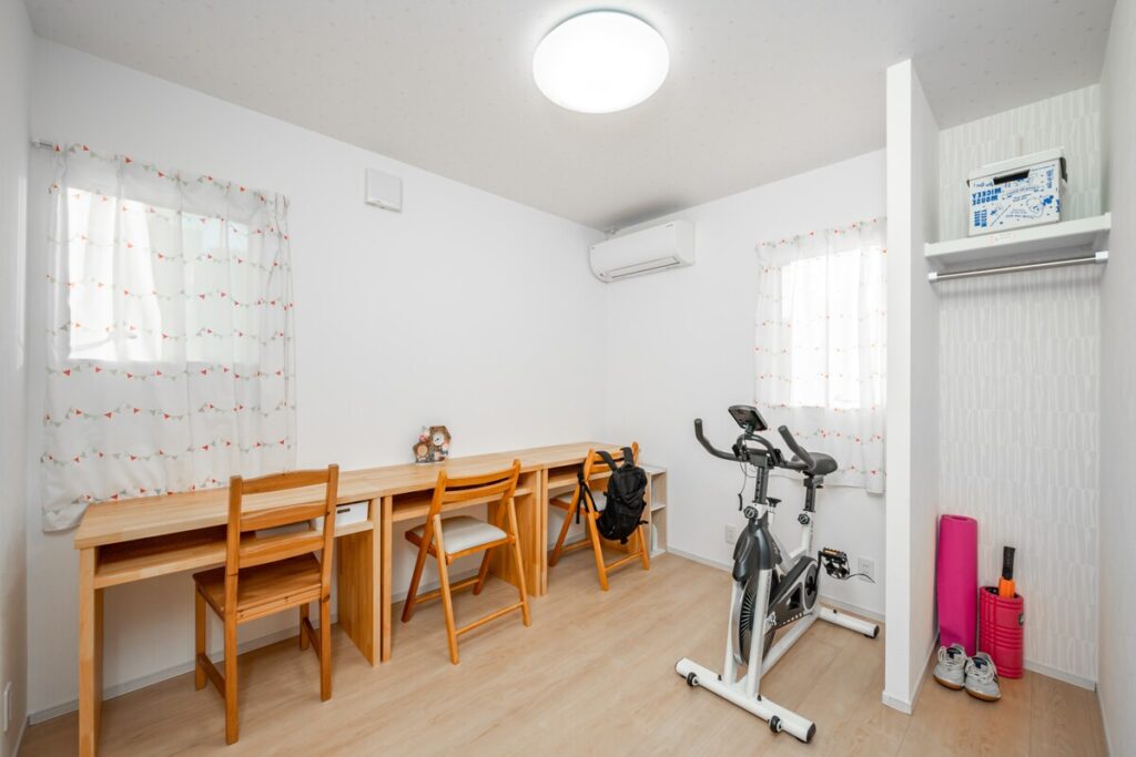 3人並んで使える造作のロングデスクとフィットネスバイクを配置した、多目的に活用できる明るい洋室。
