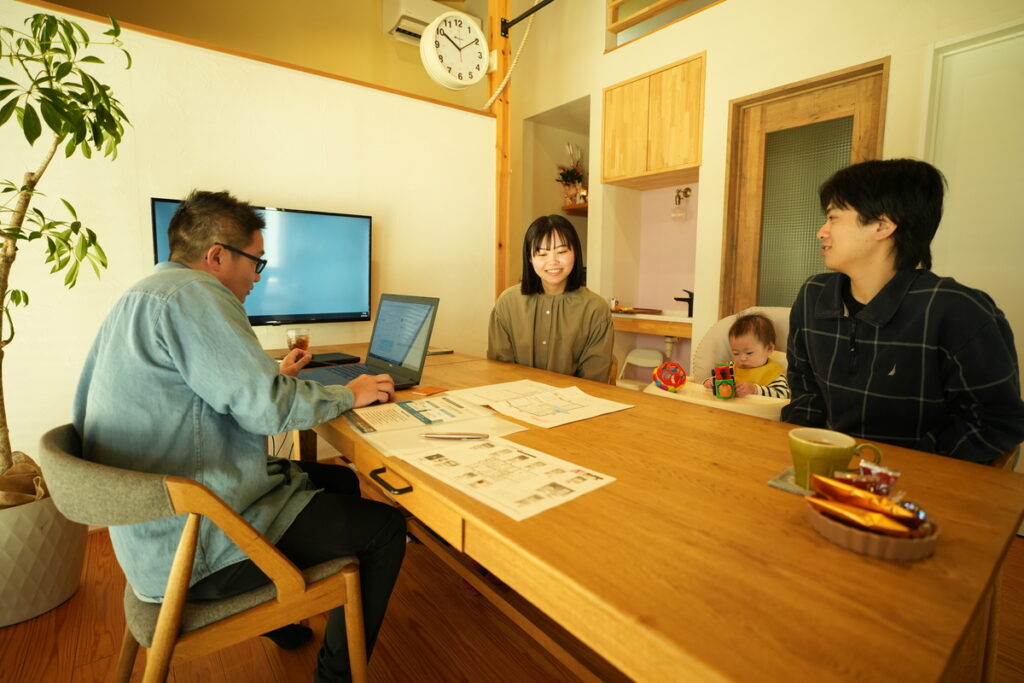 木の温もりあふれる打ち合わせスペースで、ノートパソコンや図面を広げながら、工務店のスタッフとリラックスした様子で家づくりの相談をする子連れの若い夫婦。