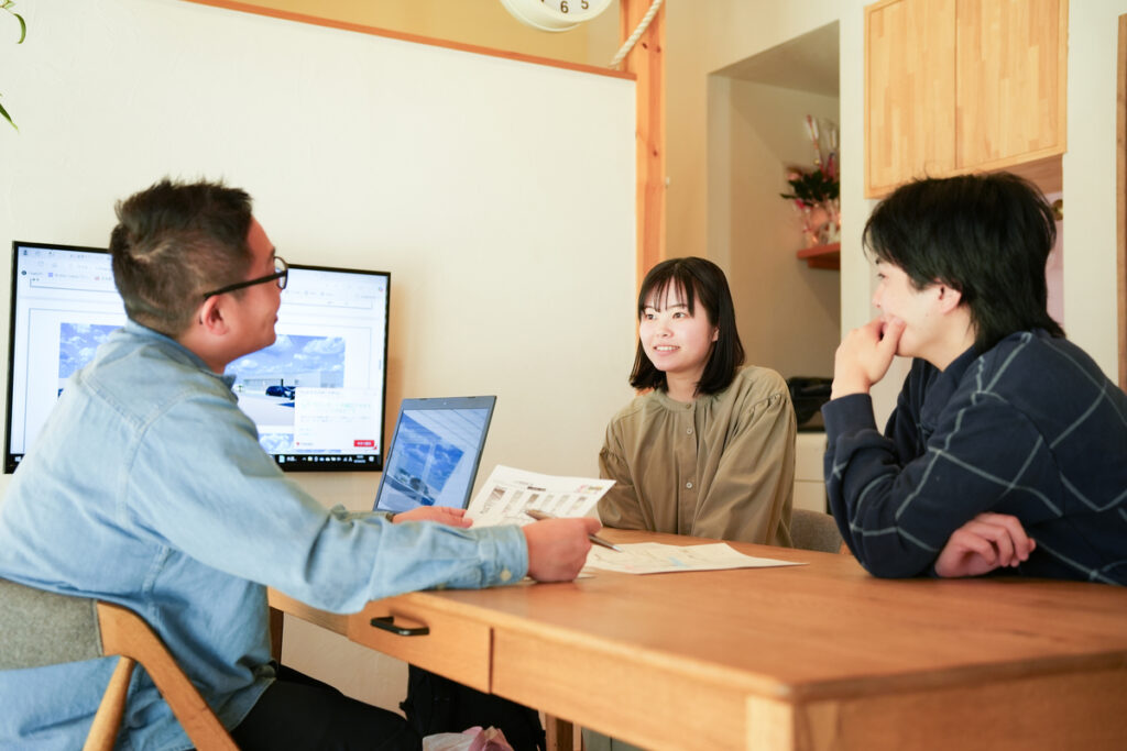 木の温もりあふれる打ち合わせスペースで、資料を手に取りながら工務店スタッフの説明を真剣な表情で聞く、家づくりを検討中の若い夫婦。