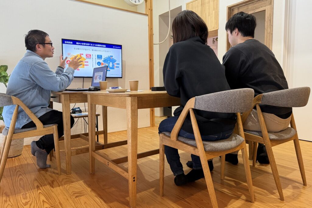 HIKAGEの打ち合わせスペースにて、モニターに映し出された住宅性能の資料を見ながら打ち合わせを行うスタッフとお客様。冬暖かく夏涼しい住まいの仕組みについて、図解を用いて分かりやすく説明している個別相談の様子です。