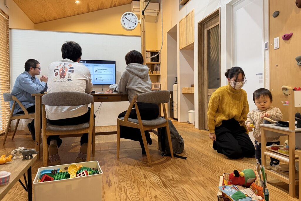 工務店の打ち合わせスペースで、モニターを見ながら家づくりの相談をする家族とスタッフ。木の温もりあふれる室内にはキッズスペースが併設されており、子供を遊ばせながらリラックスして相談できる環境が整っています。