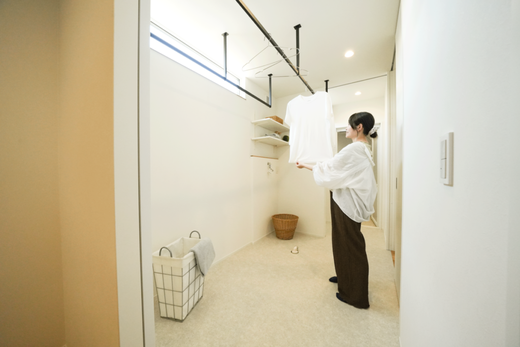 ホワイトで統一された清潔感のあるランドリールーム。天井付けの黒いアイアンハンガーパイプに洗濯物を干す様子や、可動棚による収納スペースを備えた家事効率の良い室内干し空間です。