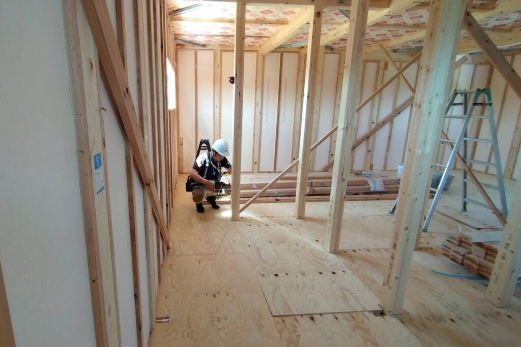 木造住宅の建築現場で、ヘルメットを着用した施工スタッフが床下に潜り、構造体や断熱材の状態を細かくチェックしている品質管理の様子。