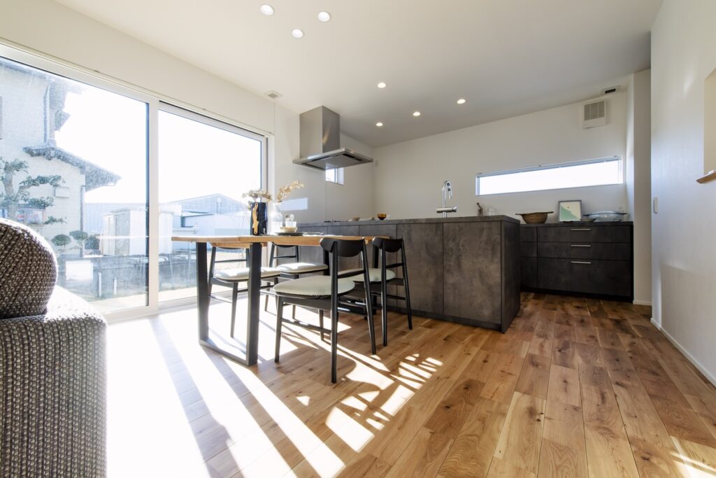 高級感のあるダークトーンのキッチンカウンターと、木の温もりを感じるダイニングセットが並ぶLDK。大きな掃き出し窓から自然光が広がり、シックなデザインとナチュラルな素材が融合した上質な空間を演出しています。