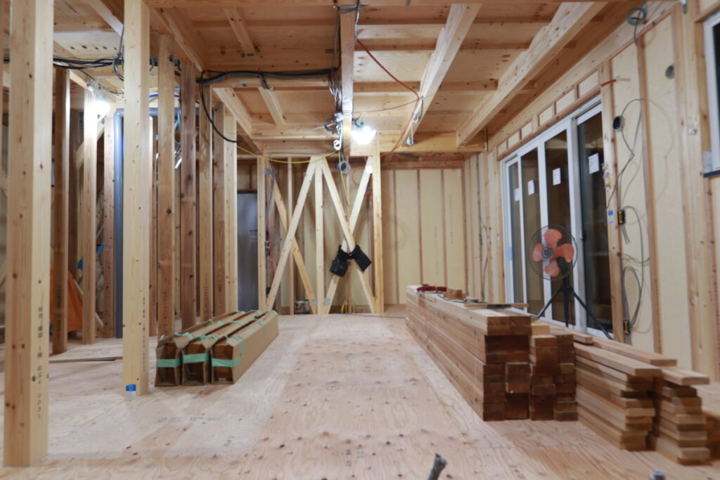 木造住宅の建設途中の構造躯体。等間隔に並ぶ柱や梁、壁面の筋交いなどが確認でき、天井裏を走る配線や断熱材の施工前の様子を捉えた現場写真です。