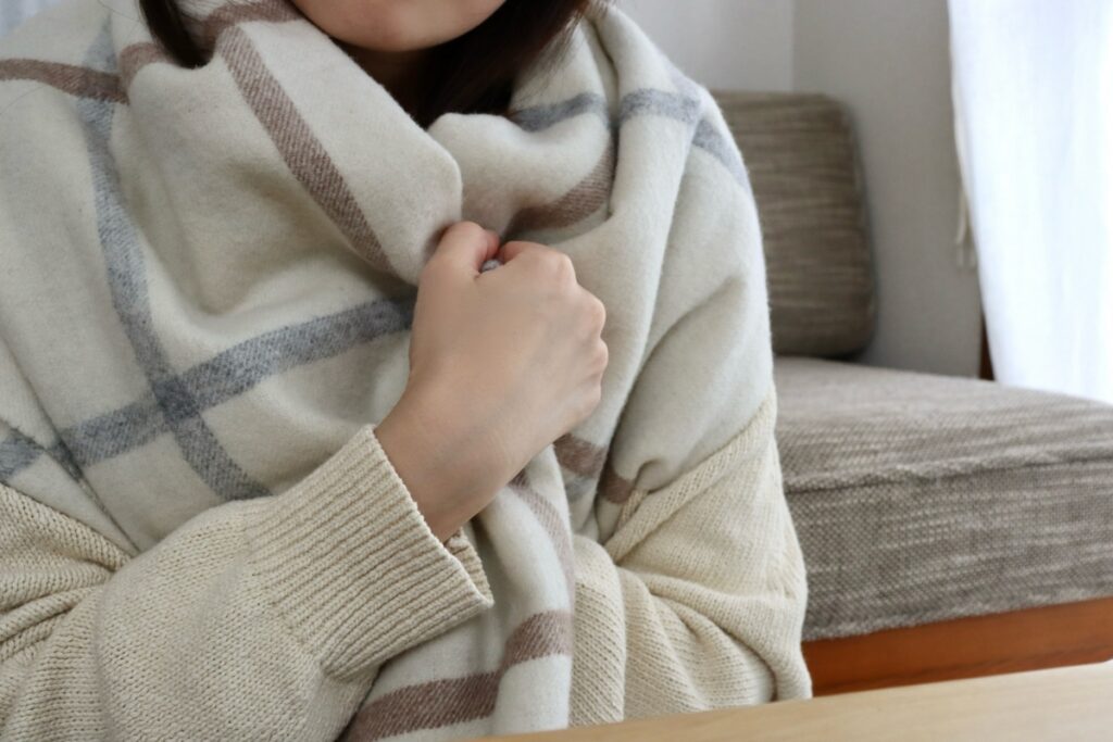 寒い室内で厚手のストールを羽織り、肩をすくめて寒さに耐える女性の様子。