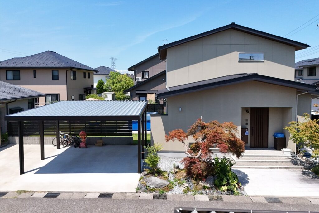 グレーの外壁としころ屋根が特徴的な和モダンの外観。手前には積雪に強い頑丈な折板屋根のカーポートと、美しく整えられたシンボルツリーのある外構が広がっています。