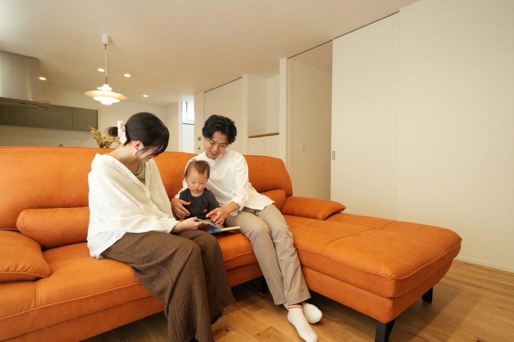 鮮やかなオレンジ色のソファに座り、お子様と一緒に絵本を楽しむご家族。無垢の床や白い建具に囲まれた明るいリビングで、家族の絆が深まる穏やかな日常のひとときを映し出しています。