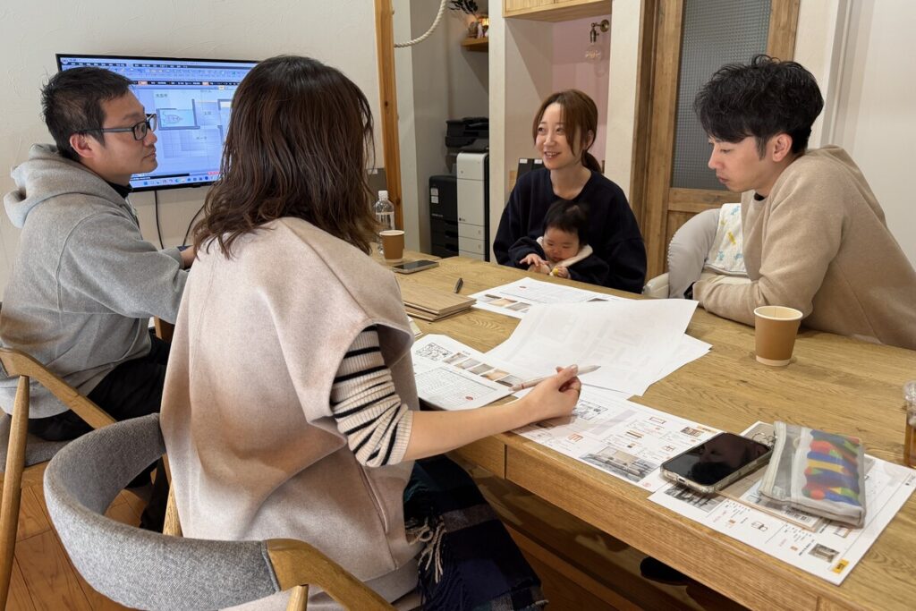 モニターの間取り図を指しながら、赤ちゃんを連れた家族と工務店スタッフがプランの細部を相談している家づくりの打ち合わせ。
