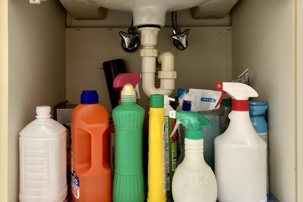 洗面台下の収納内に、排水パイプを囲むようにして乱雑に詰め込まれた色とりどりの洗剤ボトルや掃除用具。