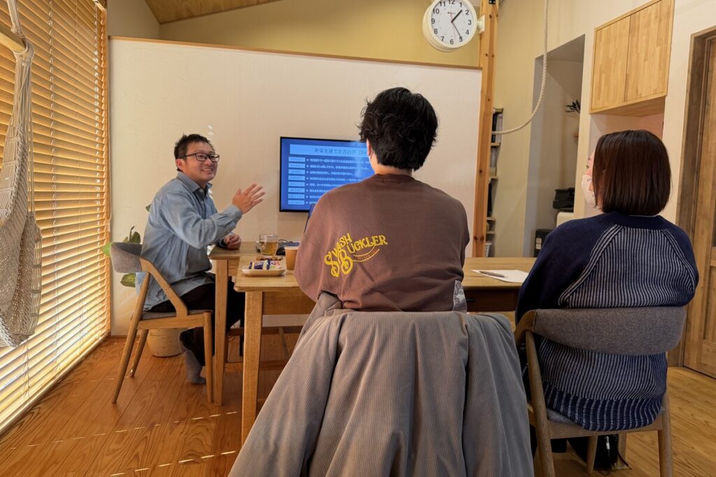 モニターに映る「新築を建てた方の声」を見ながら、工務店のスタッフから家づくりのアドバイスを受ける夫婦の打ち合わせ風景。